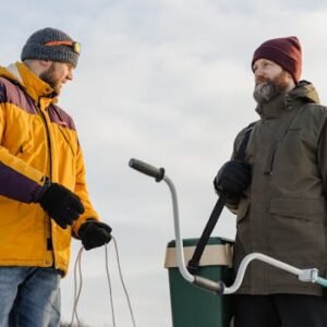 Two bearded men in winter clothes ice fishing outdoors with equipment.